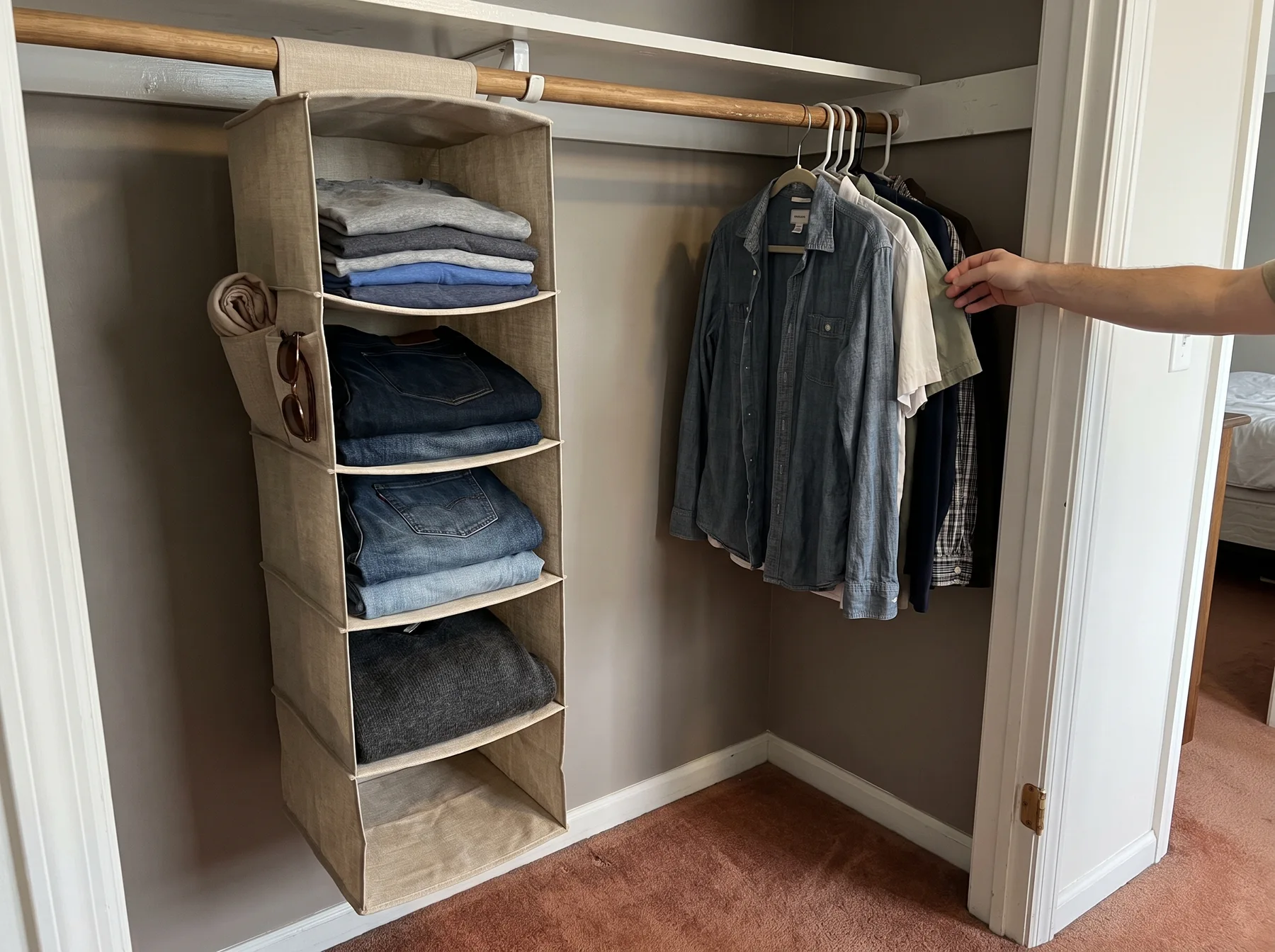 Fabric shelf organizer holds folded clothes beside shirts hanging from a closet rod.