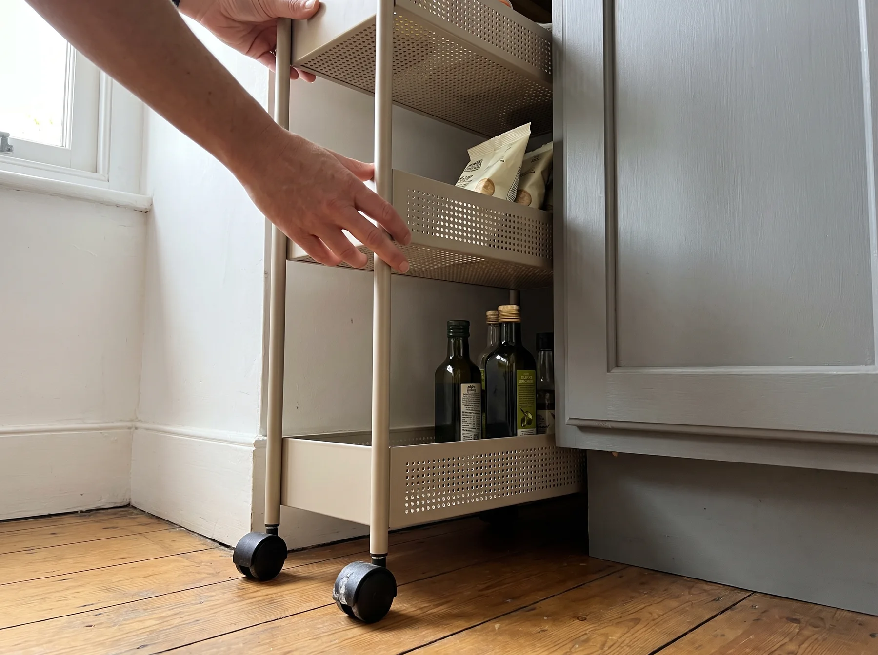 Hands pull a narrow rolling storage cart from beside a kitchen cabinet.