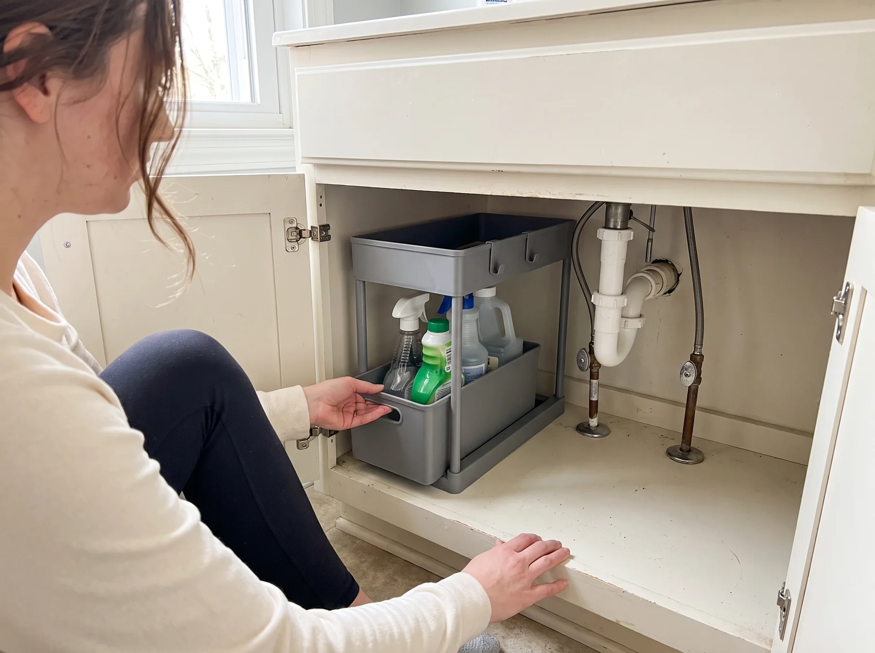 Woman pulls out a tiered organizer beside exposed pipes under a bathroom sink.
