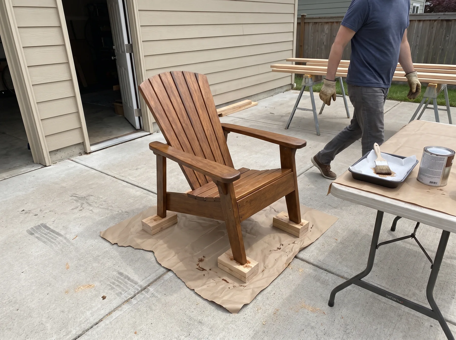 Wooden fan-back Adirondack chair on blocks in a driveway beside a table with finishing supplies