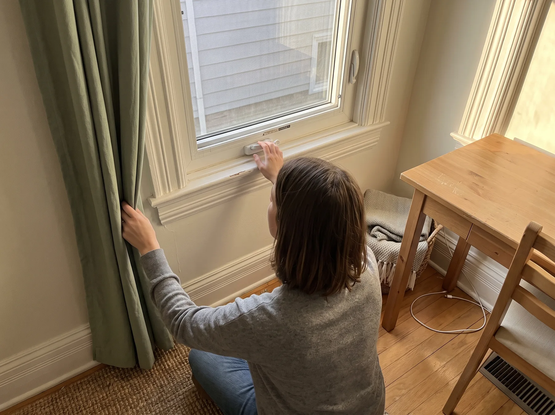 Woman kneels beside a bedroom window, checking the latch while holding a curtain near an unobstructed desk.