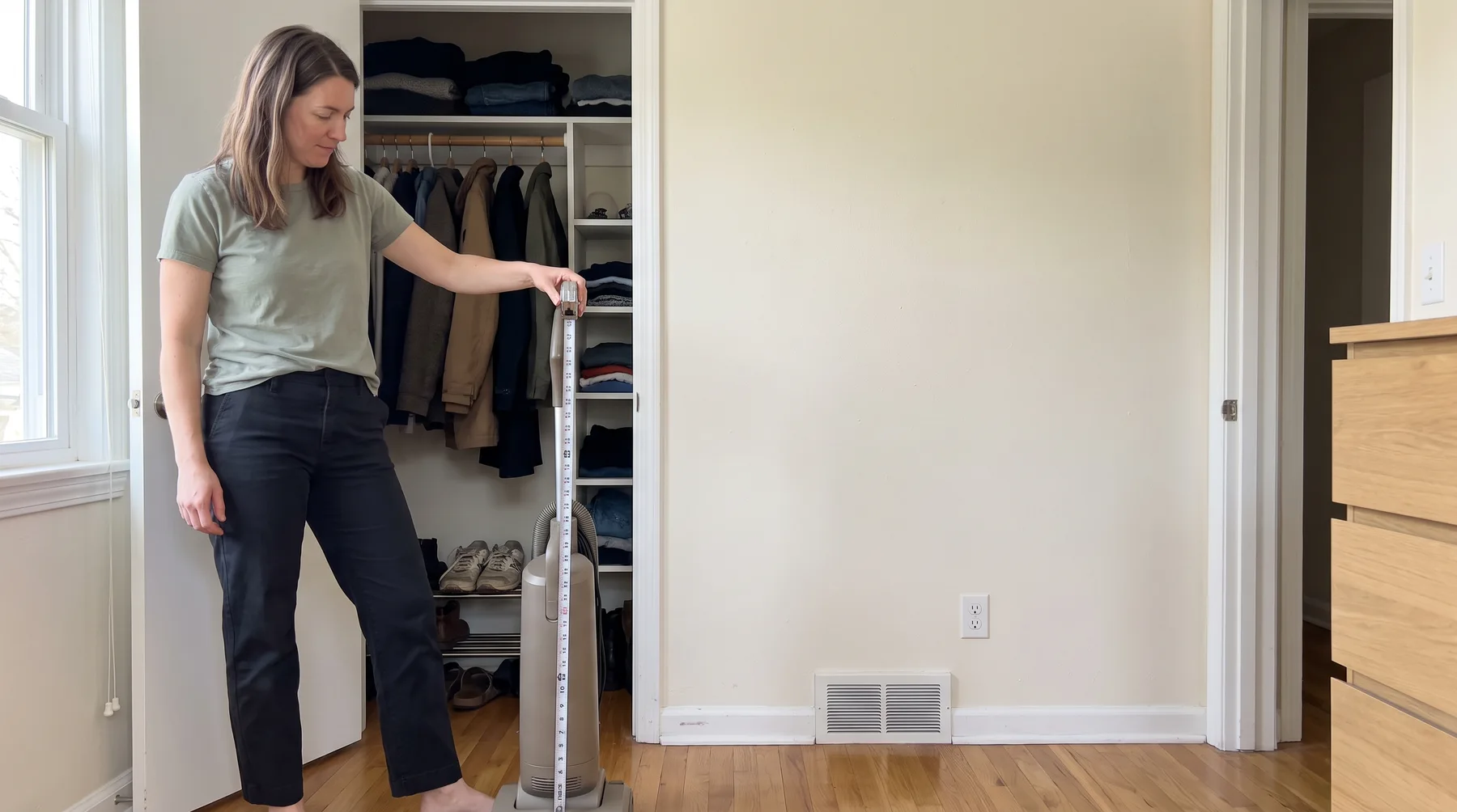 Woman measures an upright vacuum beside an open closet in a small apartment.