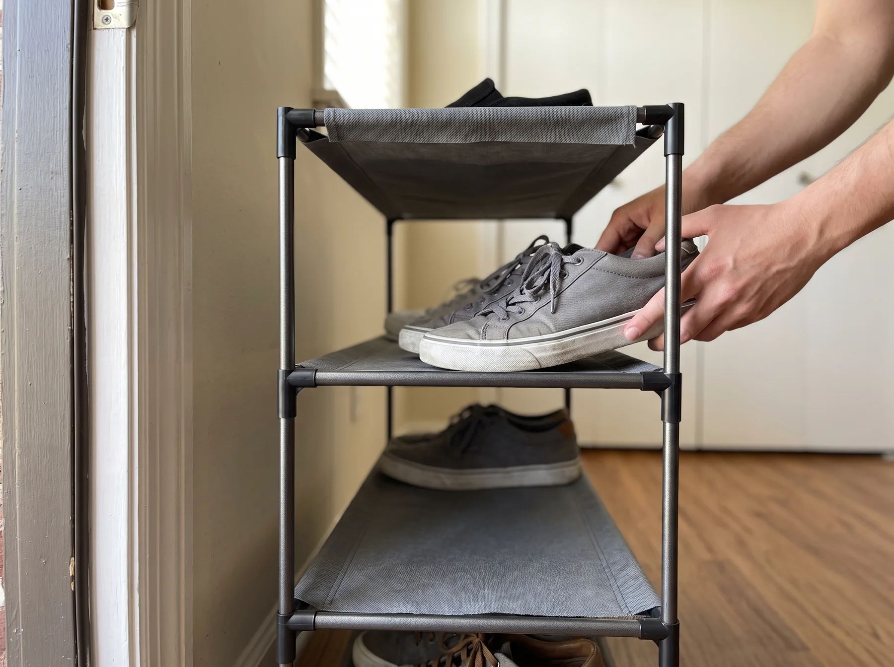Hands place gray sneakers on a narrow shoe rack inside an apartment doorway.