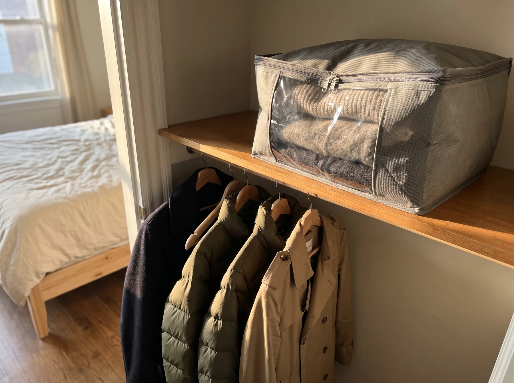 Mesh storage bag sits above hanging coats in a bedroom closet beside a bed.
