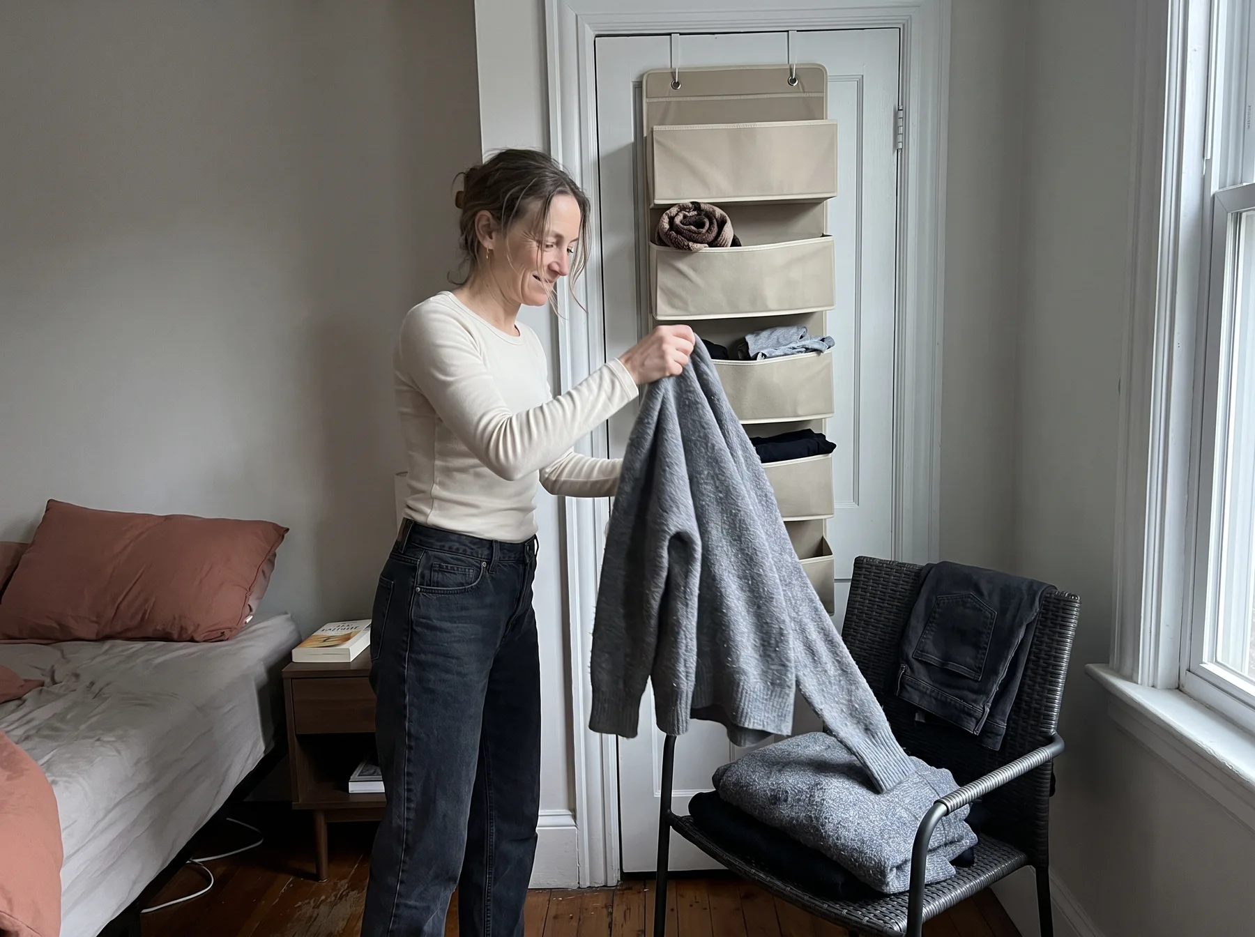 Woman holds a gray sweater beside a chair and hanging organizer in a small bedroom.