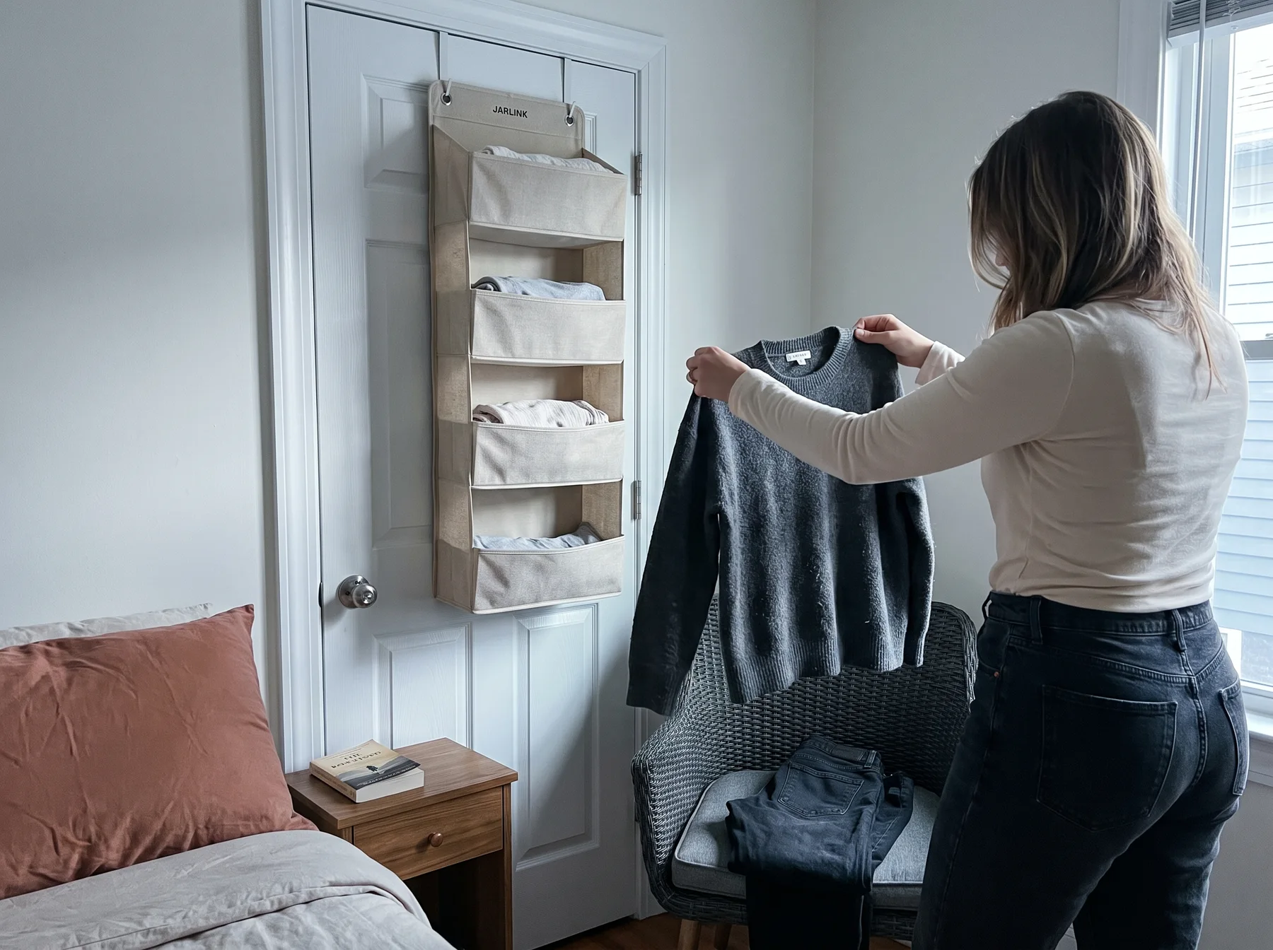 Woman lifts sweater from clothing-covered chair beside an over-door organizer in bedroom.